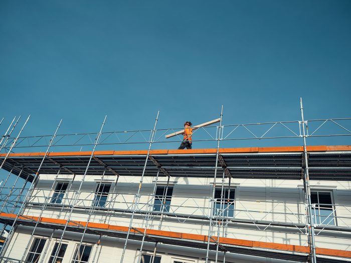 En byggarbetare som bär på planka gåendes på byggnadsställning