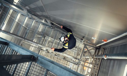 Man som klättrar i trappa på hög höjd. Industriarbetaren bär skyddshjälm.